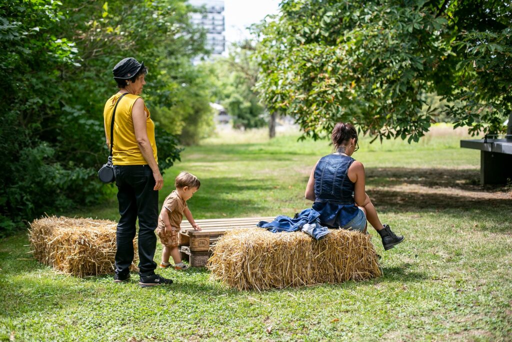 A woman sitting on a bale of straw, next to her a small child and an elderly lady in a yellow T-shirt.